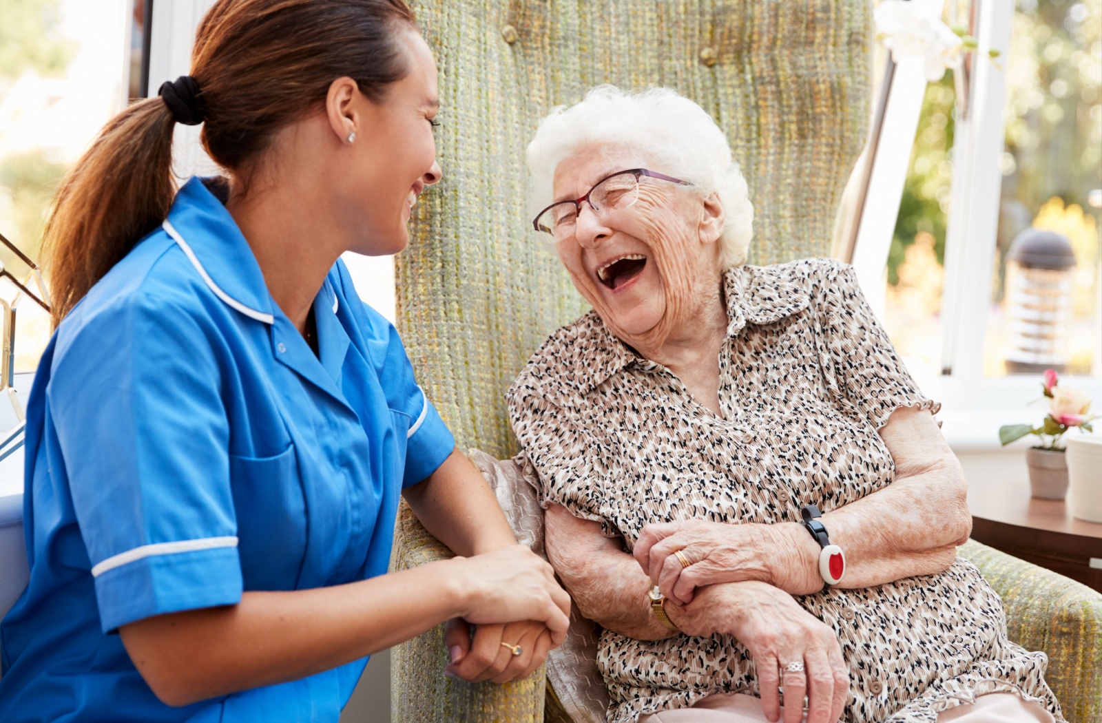 An older woman sitting in a chair laughs with her caretaker professional.