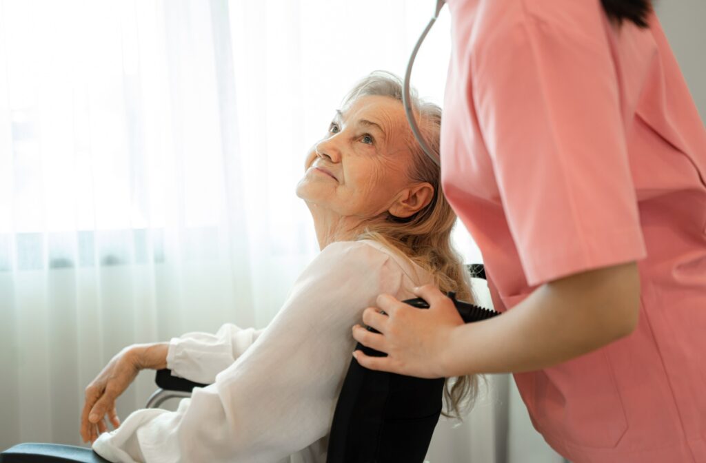 An elderly woman seated in a wheel-chair looks warmly up at a woman in pink scrubs.