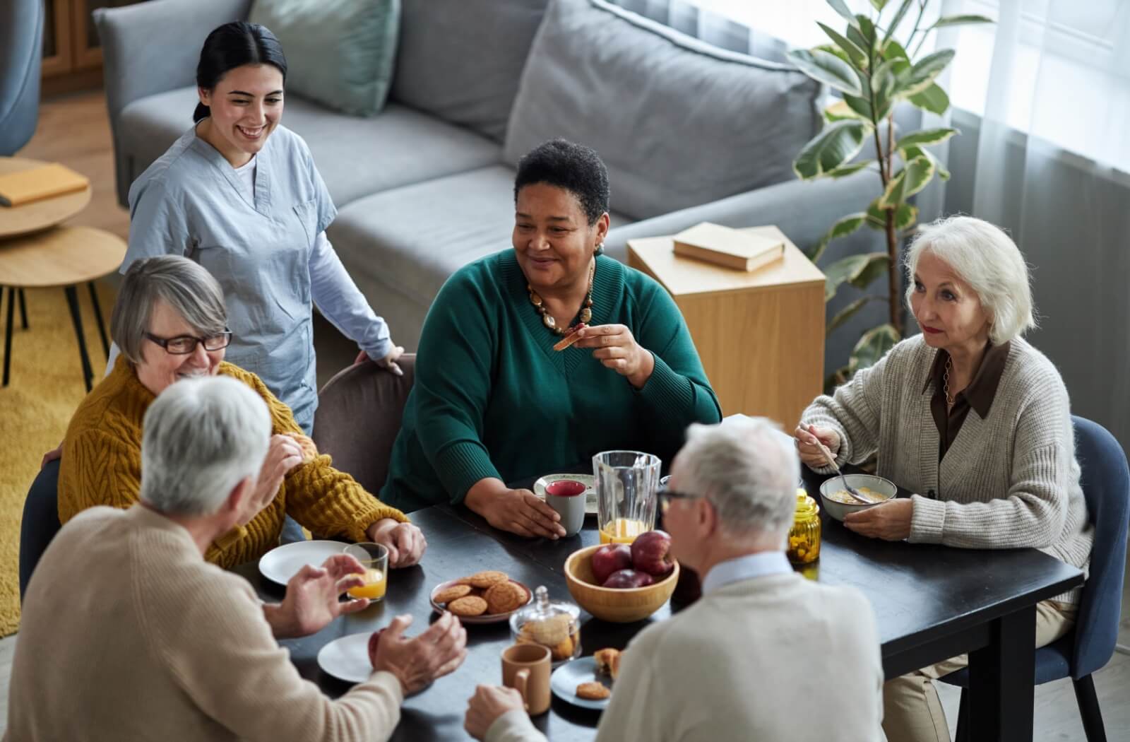 A group of seniors chat over breakfast at an assisted living community while a nurse checks in with them.