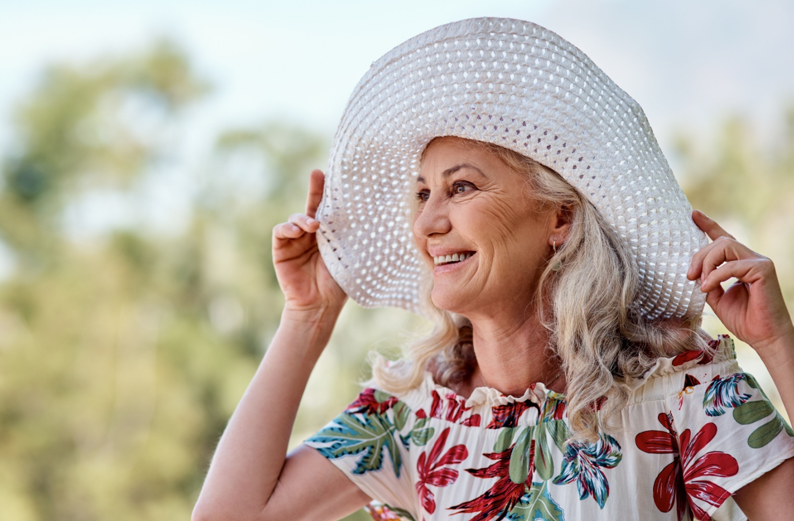 Smiling older woman wearing a wide-brimmed sun hat and floral dress outdoors on a sunny day, staying cool and protected from the summer heat.