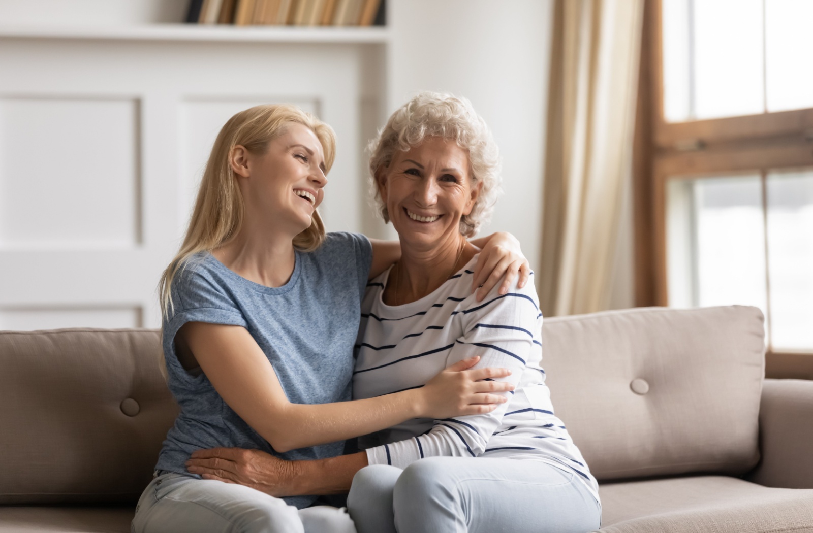 Adult daughter and senior mother sitting on a couch, smiling and embracing during a relaxed visit in an assisted living community.