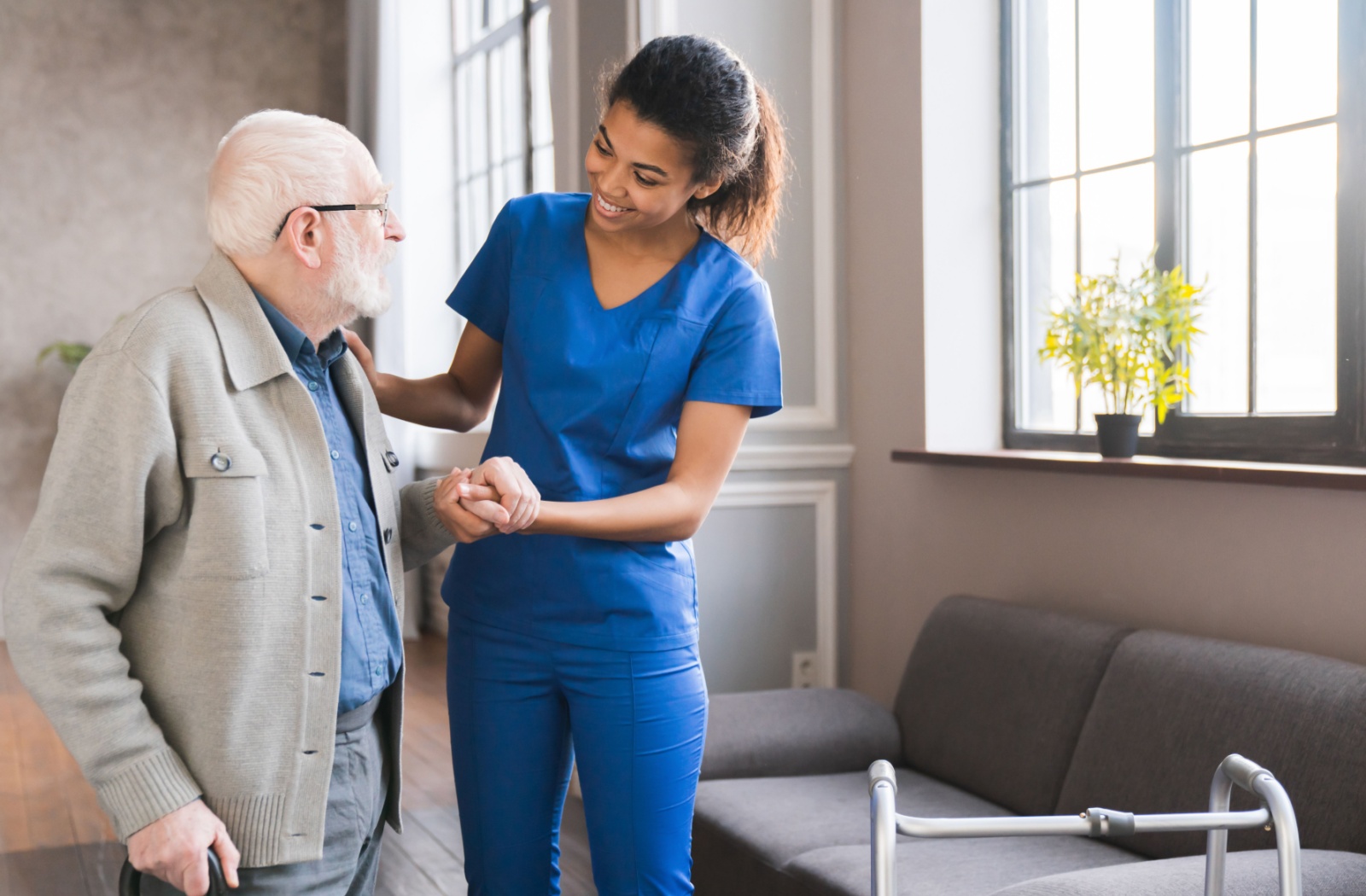A nurse in blue scrubs helping a senior with a walker at an assisted living community.