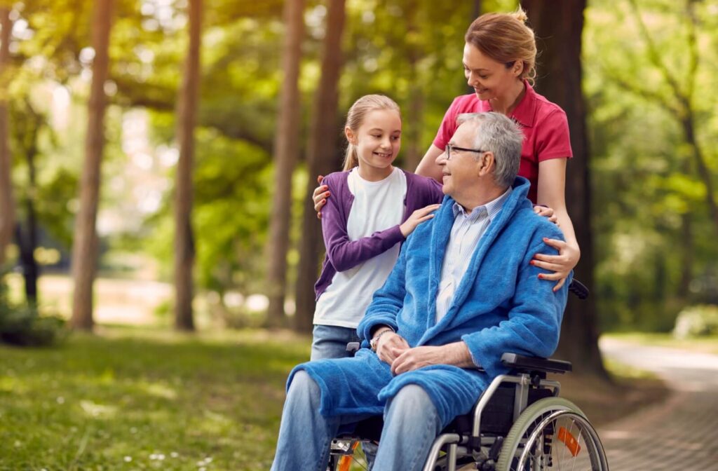 Senior man in a wheelchair spending time outdoors with his daughter and granddaughter during a family visit at an assisted living community.