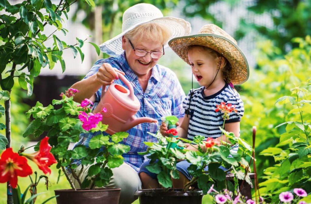 Older woman and young girl wearing sun hats while watering flowers together in a garden, enjoying a safe and sunny summer day outdoors.