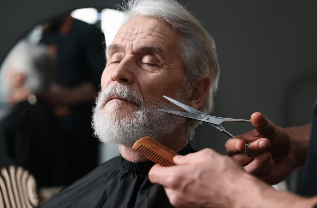 A senior closes their eyes while a professional barber trims their beard, a unique service offered in assisted living