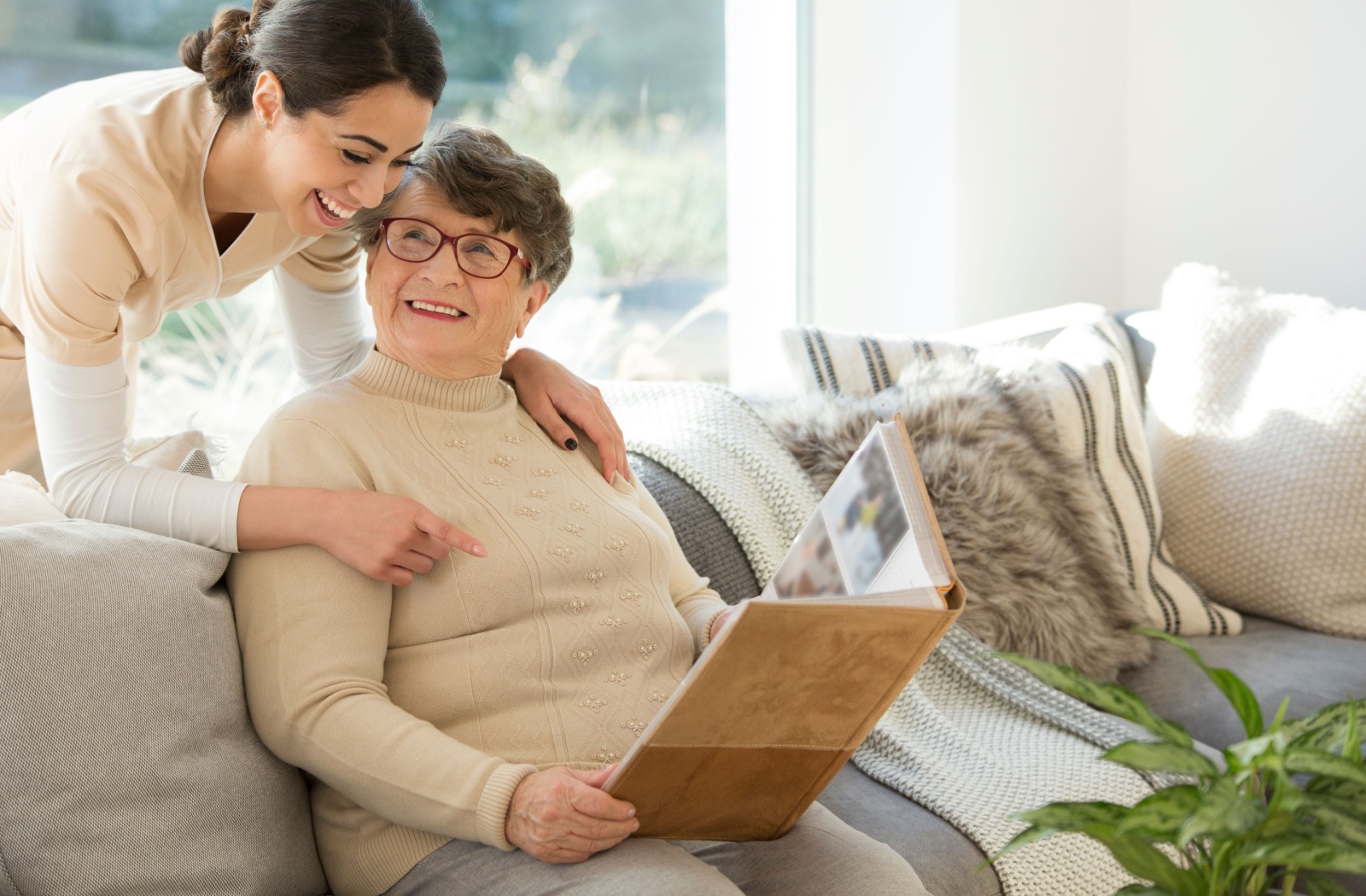 An adult leans over the couch to hug their senior parent while looking at a photo album.